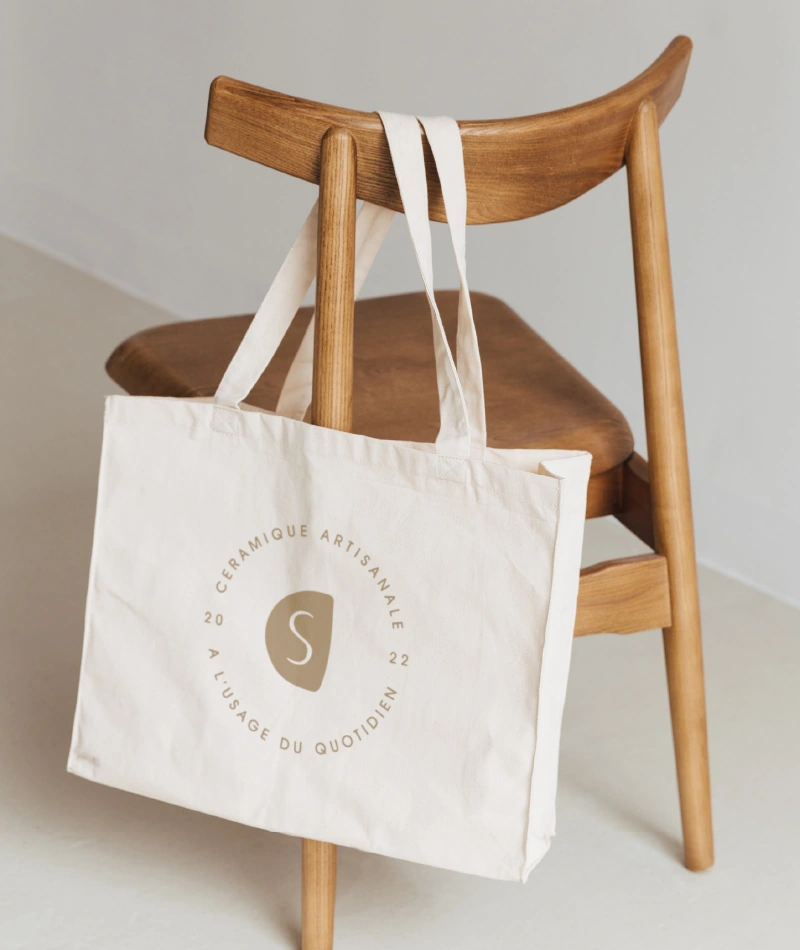 Un totebag large est accroché à une chaise en bois miel. De couleur claire, il comporte le macaron de l'identité de marque Claire Schott, qui porte le texte "Céramique artisanale, à l'usage du quotidien, 2022".